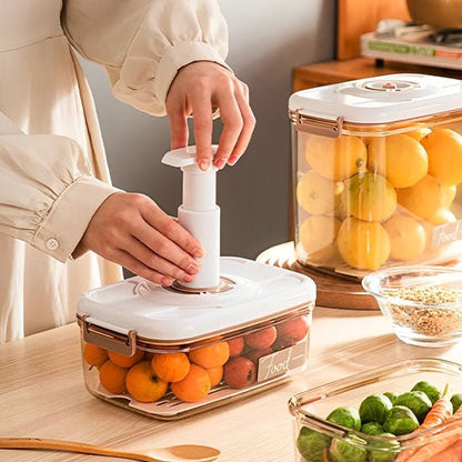 Boîte de conservation alimentaire sous vide avec piston et fruits à l’intérieur sur plan de travail, entourée d’autres contenants et légumes.