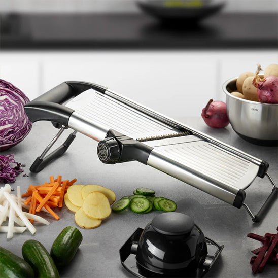 Mandoline cuisine réglable noire et argent avec lames; légumes tranchés autour et bol de légumes au fond, plan de travail moderne.