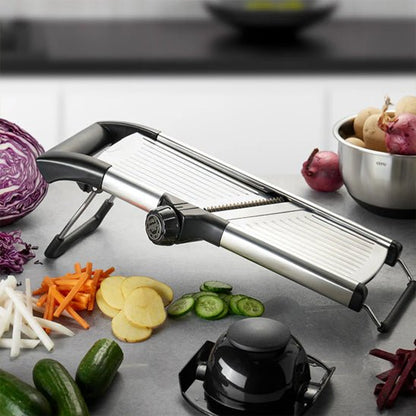 Mandoline cuisine réglable noire et argent avec lames; légumes tranchés autour et bol de légumes au fond, plan de travail moderne.